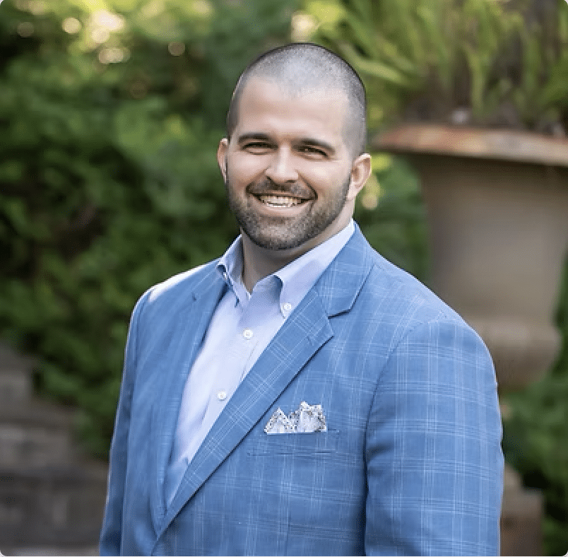 A smiling man with a shaved head and beard wears a light blue plaid suit jacket and a white dress shirt, standing outdoors in front of greenery and stone steps.